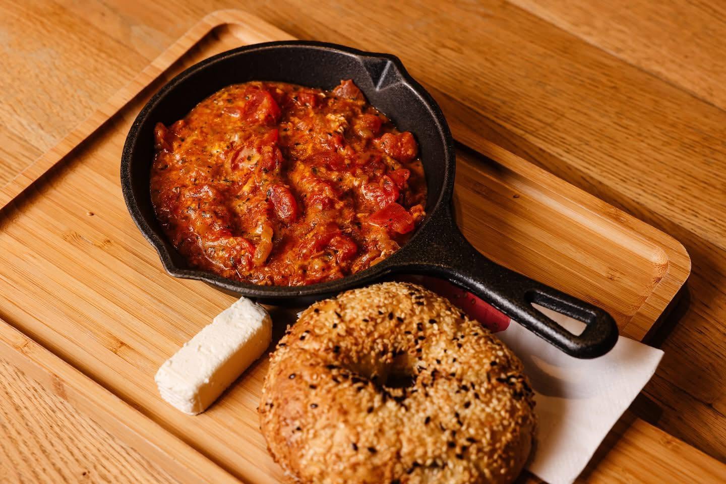 Menemen në tigan gize me bagel me susam dhe krem djathi, mbi tryezën e drurit të Fikës